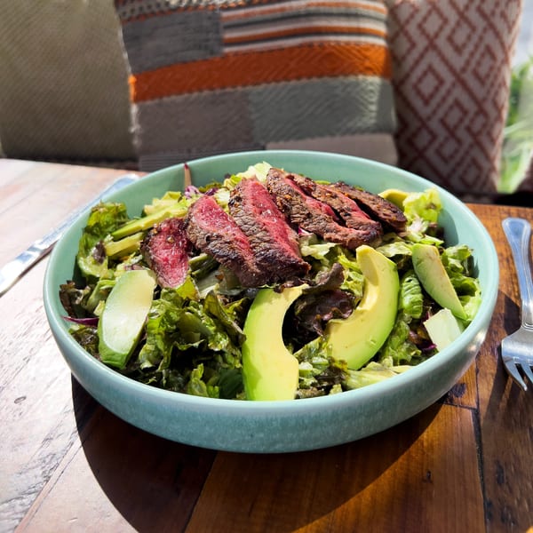 A vibrant steak and avocado salad in a teal bowl on a wooden table at Juicy Ladies.