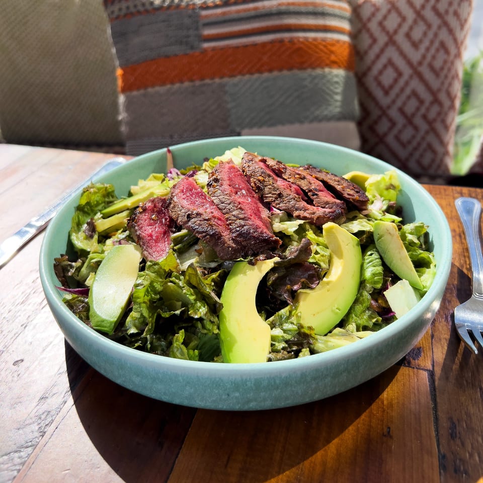 A vibrant steak and avocado salad in a teal bowl on a wooden table at Juicy Ladies.
