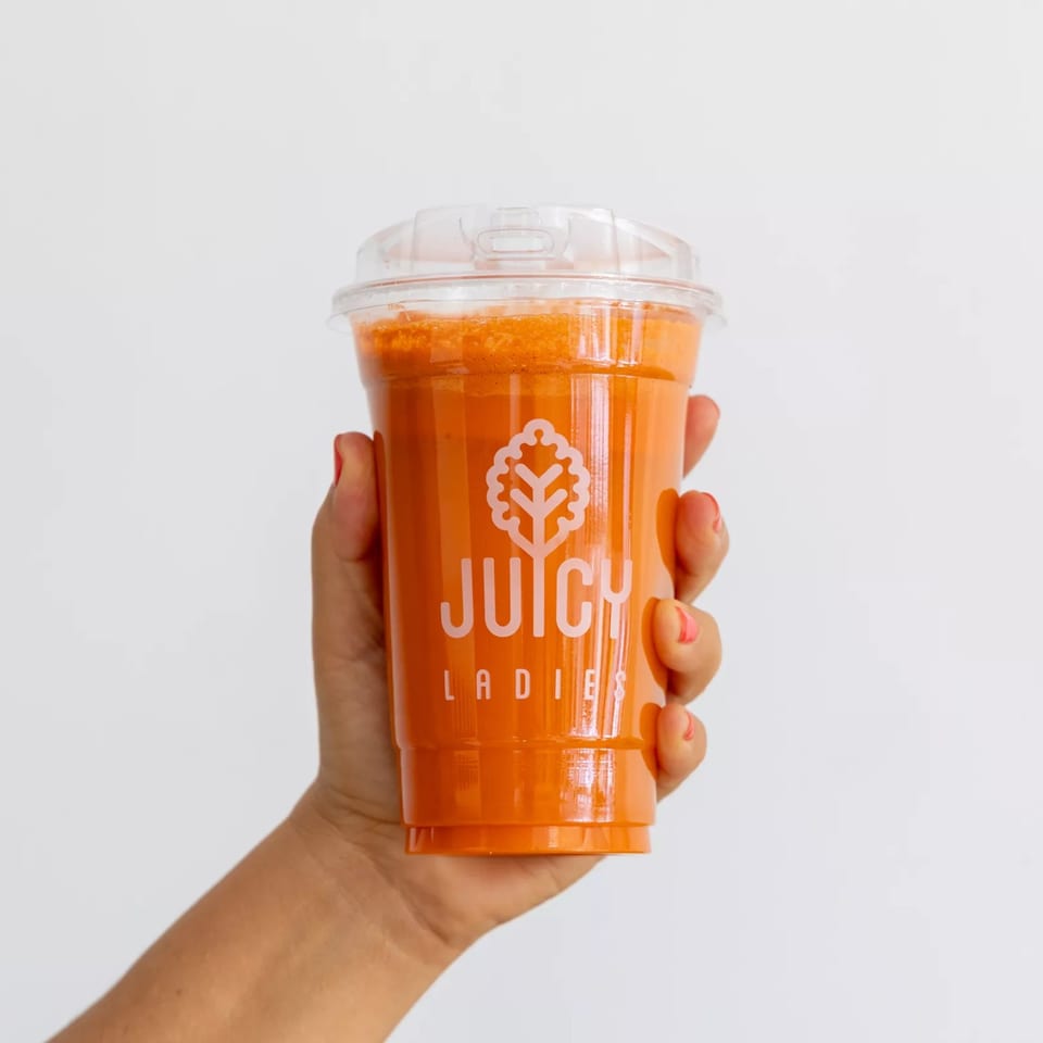 Hand holding a clear JUICY LADIES cup filled with bright orange juice against a plain white background.