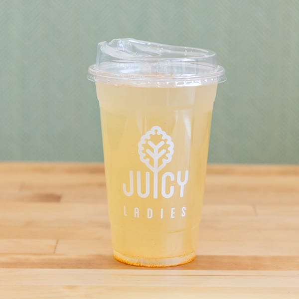 Yellow beverage in a clear plastic cup labeled Juicy Ladies, resting on a wooden table against a green background.