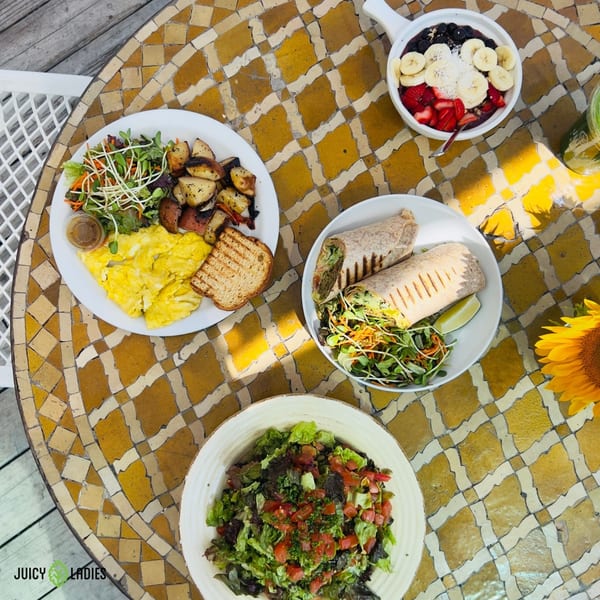 Overhead view of a mosaic table laden with healthy dishes from Juicy Ladies, including breakfast, wraps, and açaí bowl.