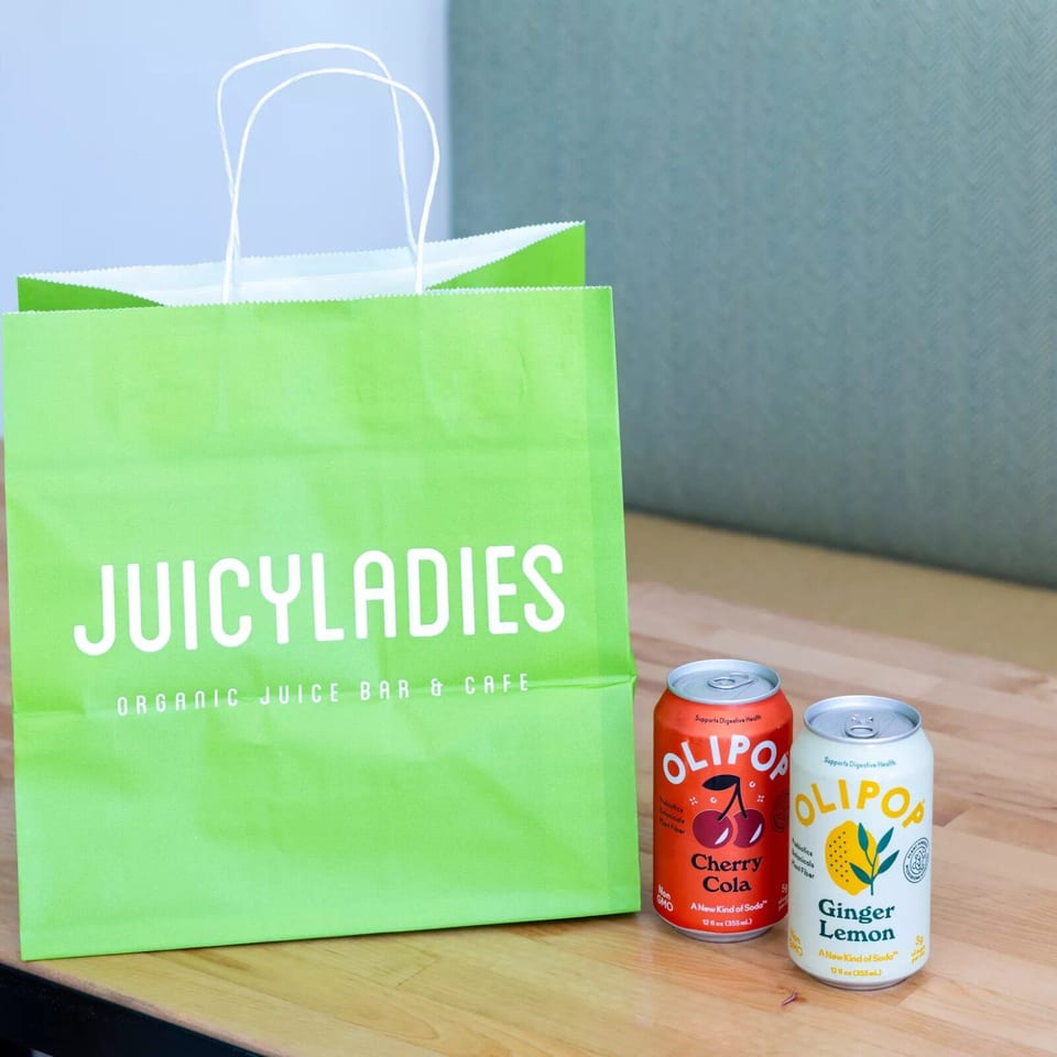 Green Juicyladies organic juice bar bag beside cans of Ollipop Cherry Cola and Ginger Lemon on a wooden table.