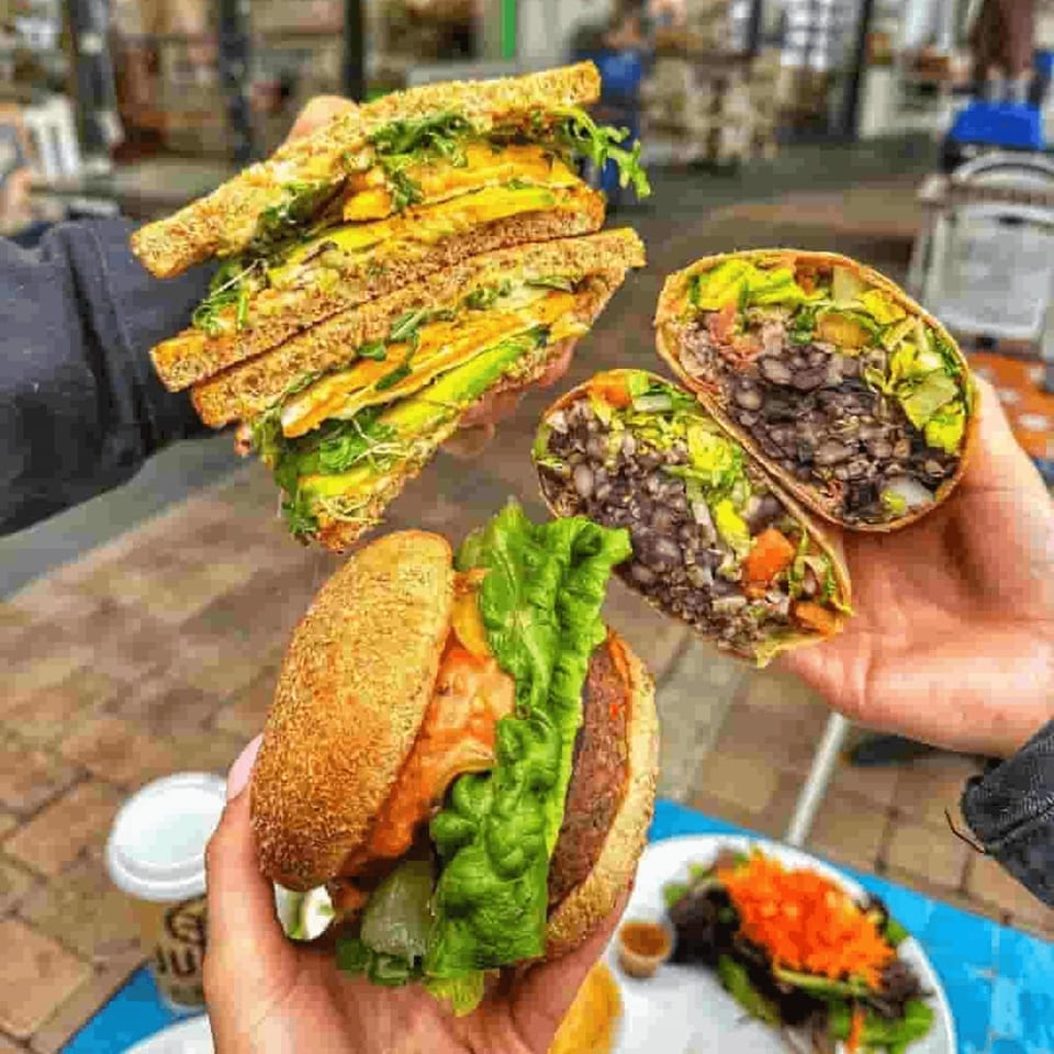 Assortment of stuffed flatbreads and sandwiches with leafy greens, vegetables, and dark beans held outdoors near a restaurant.