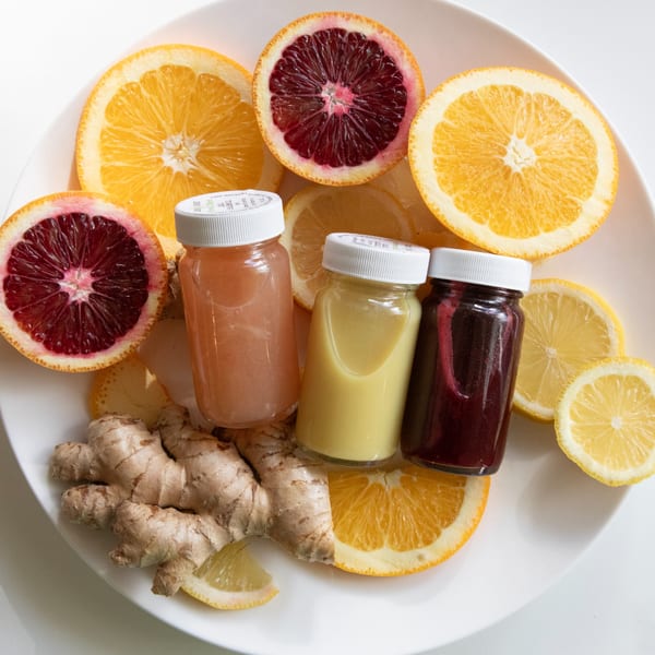 Three colorful juice shots with sliced oranges, lemons, and ginger on a white plate.