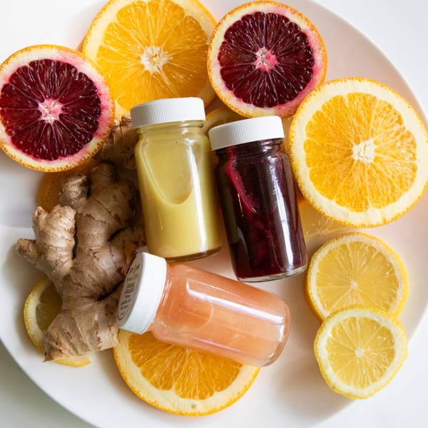 Three small bottles of colorful juices surrounded by sliced oranges, lemons, and ginger on a white plate.