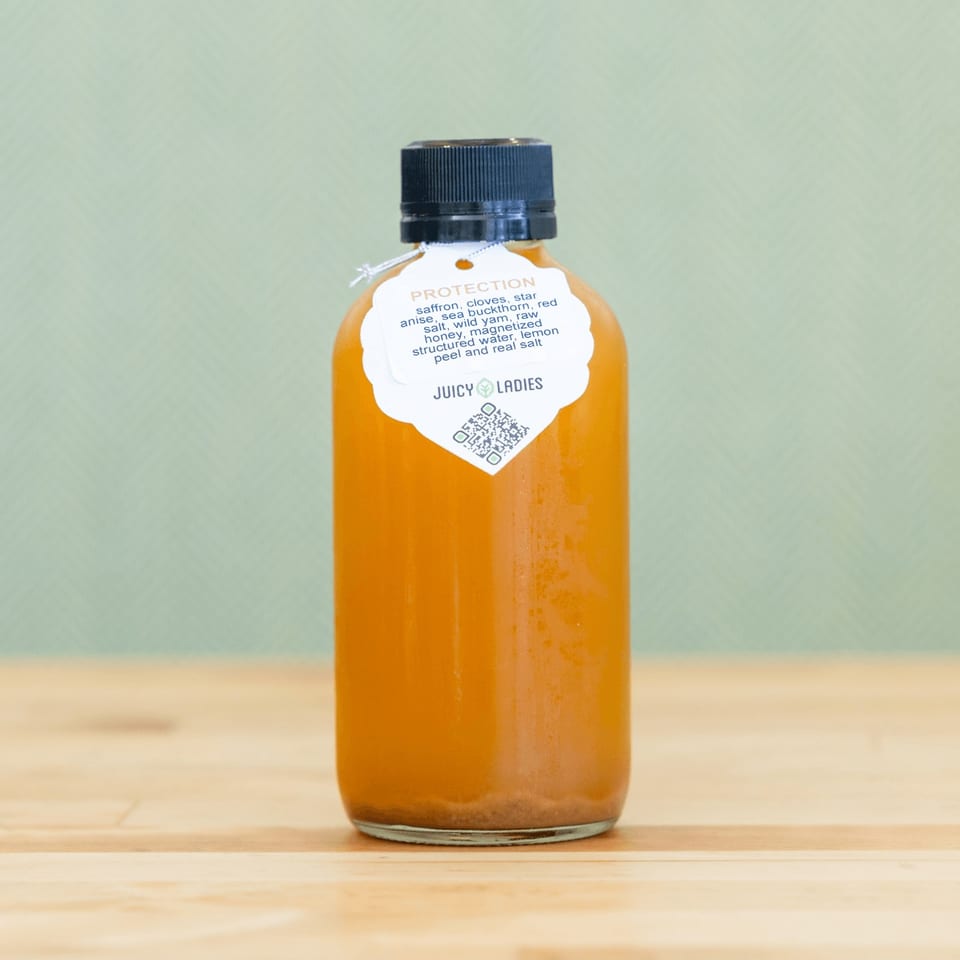 Bottle of orange juice labeled Protection by Juicy Ladies, displayed on a wooden table against a pale green background.