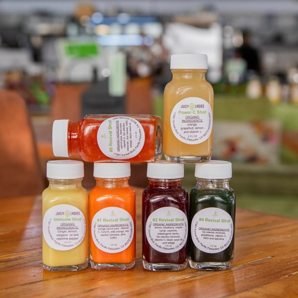 Six colorful Juicy Ladies healthy shots, including Power C, Immune, and Revival, on a wooden table.
