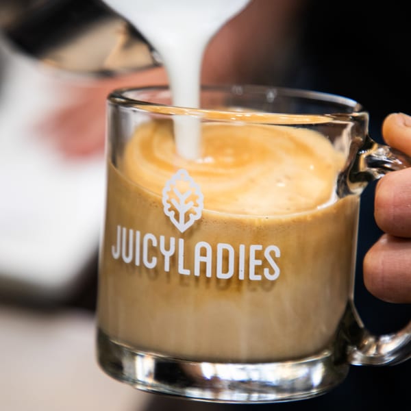 Barista pouring steamed milk into a Juicy Ladies branded glass mug of coffee.