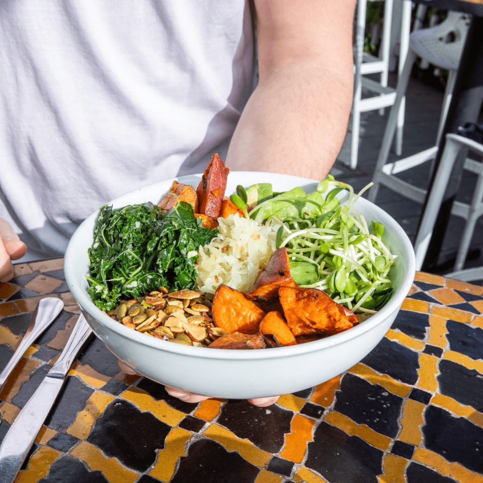 A person holds a colorful grain bowl with leafy greens, roasted sweet potatoes, seeds, rice, and vegetables over a patterned restaurant table.