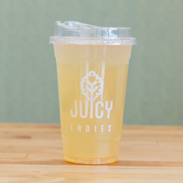Yellow drink in a clear lidded cup labeled Juicy Ladies, displayed on a wooden table against a green background.