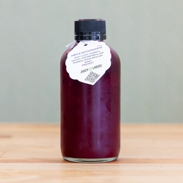 Bottle of dark red berry juice labeled Juicy Ladies Ample Antioxidants on a wooden table.