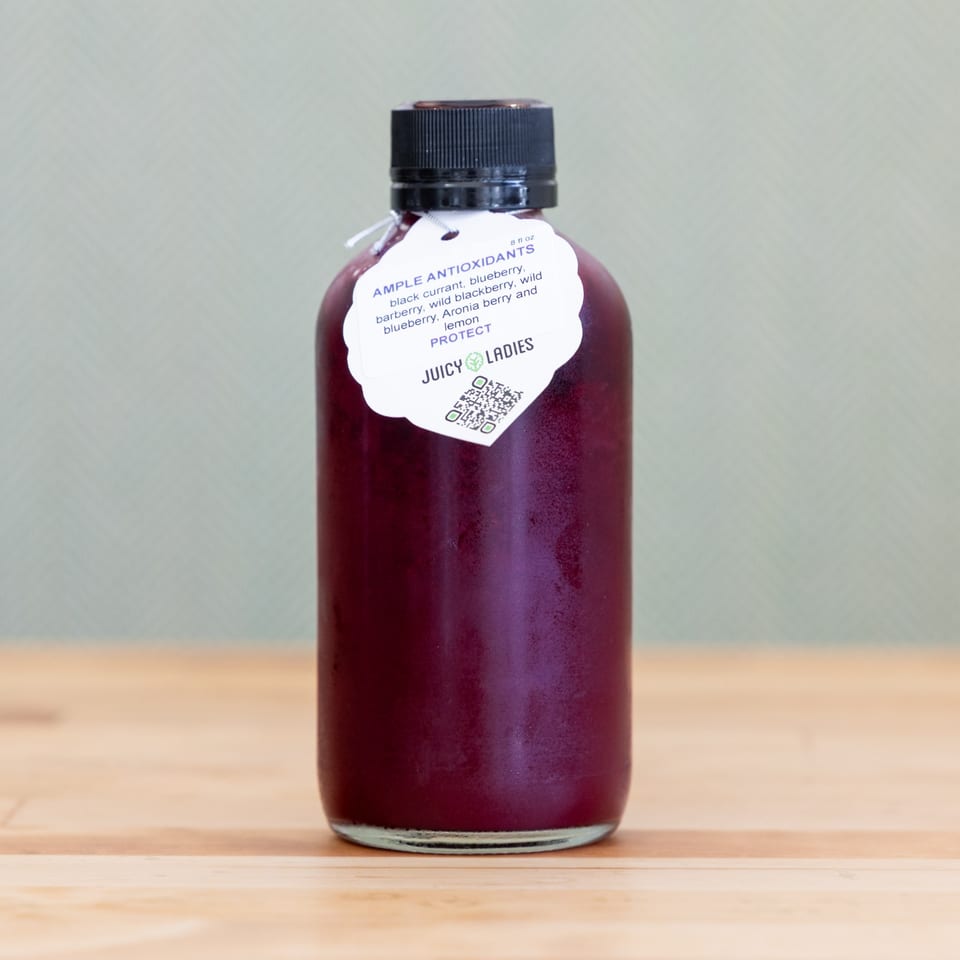 Bottle of dark red berry juice labeled Juicy Ladies Ample Antioxidants on a wooden table.