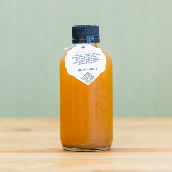 Bottle of orange-colored protection tonic labeled Juicy Ladies on a wooden table against a pale green background.