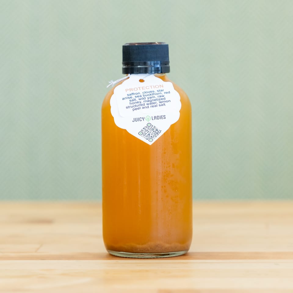Bottle of orange-colored protection tonic labeled Juicy Ladies on a wooden table against a pale green background.