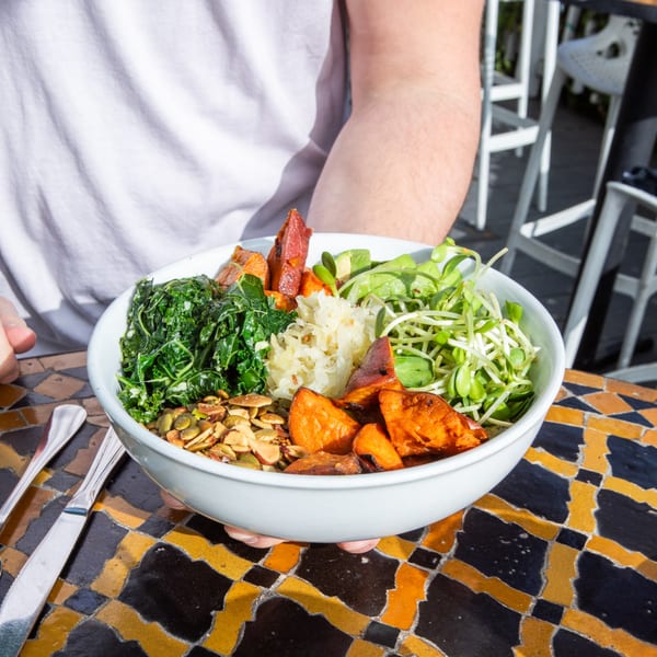 Hands holding a healthy Juicy Ladies bowl with kale, sweet potatoes, sprouts, and seeds on a tiled table.