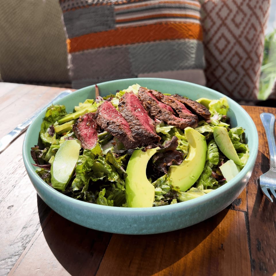 Grilled steak slices and avocado arranged over mixed greens in a turquoise bowl on a wooden restaurant table.