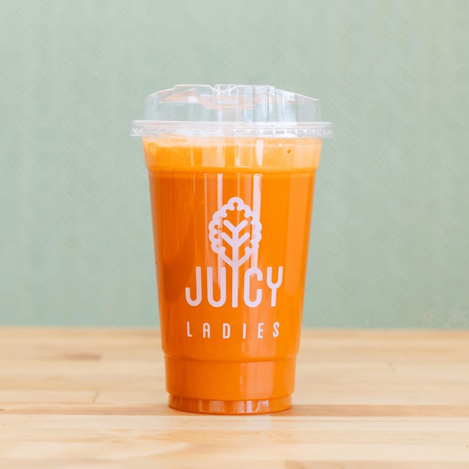 Clear takeaway cup filled with bright orange juice, branded “Juicy Ladies,” sits on a wooden table against a pale green background.