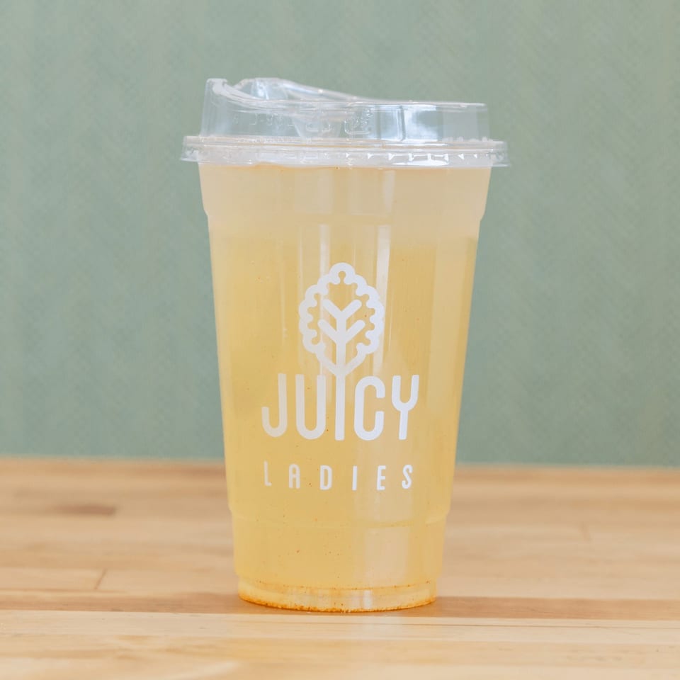 Clear cup of yellow juice labeled Juicy Ladies on a wooden table against a muted green background.