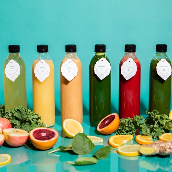 Six colorful Juicy Ladies juice bottles with fresh fruit and kale on a teal background.