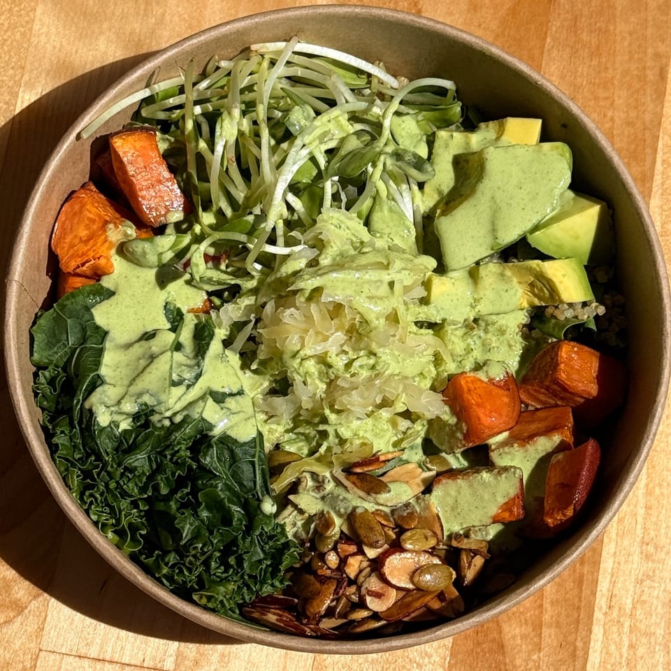 Overhead view of a colorful grain bowl with kale, roasted sweet potatoes, avocado, sprouts, nuts, and creamy green dressing.