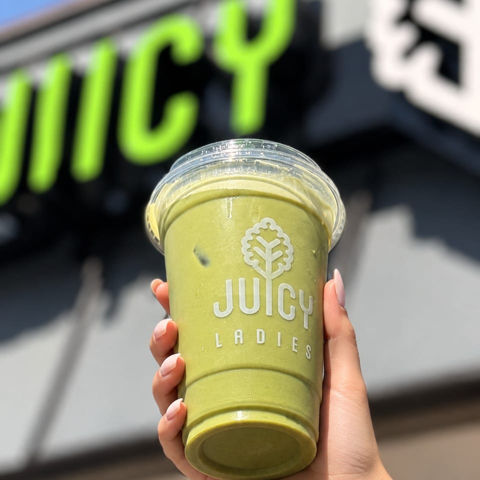Hand holding a green juice in a clear branded Juicy Ladies cup against a blurred storefront background.