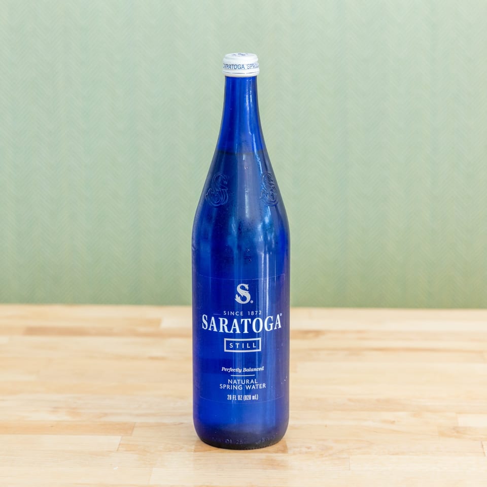 Blue Saratoga Still natural spring water bottle standing on a wooden table against a pale green background.
