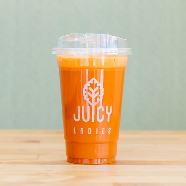Orange juice in a clear takeaway cup labeled Juicy Ladies, standing on a wooden table against a pale green background.