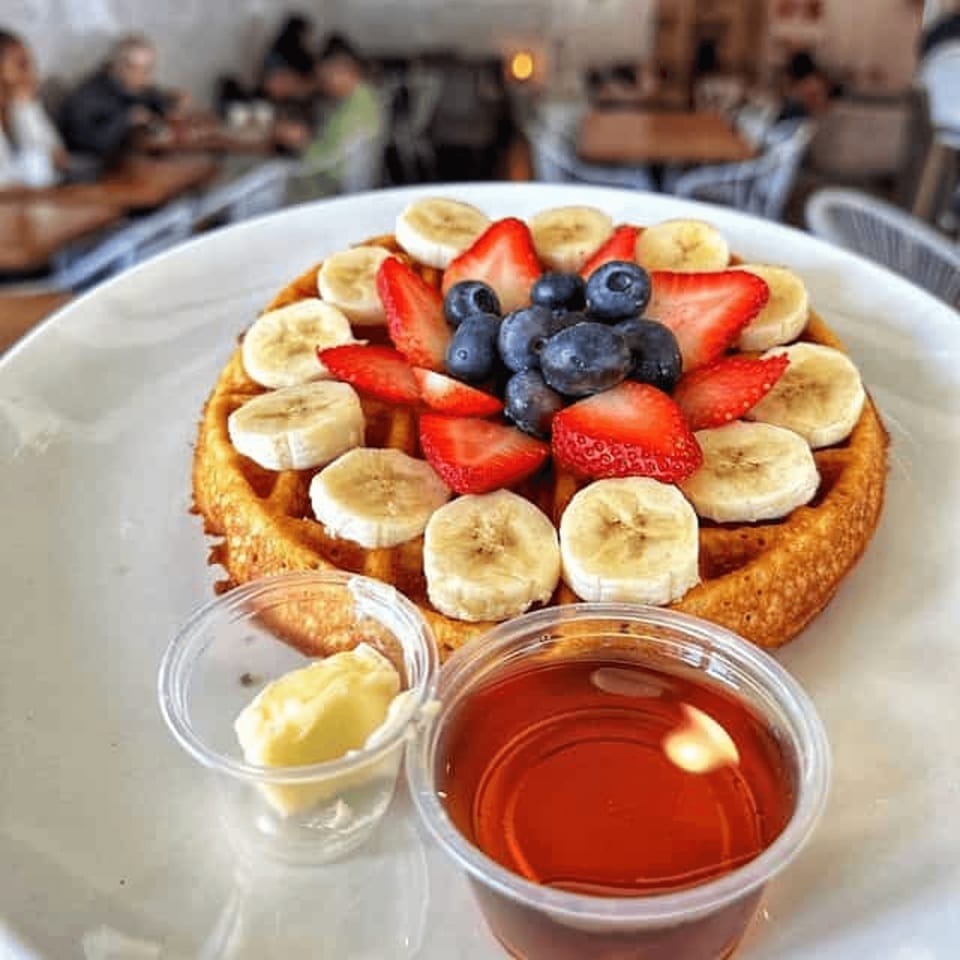 Fruit-topped waffle with strawberries, blueberries, and banana slices, served with syrup and butter in a busy café.