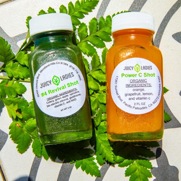 Two Juicy Ladies juice shots, green and orange, on a tiled surface with green leaves.