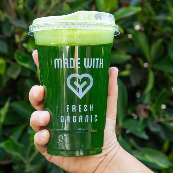 Hand holding a green juice from Juicy Ladies, with "MADE WITH FRESH ORGANIC" on the cup.