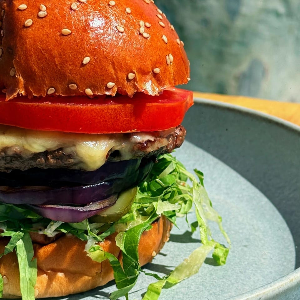 Close-up cheeseburger with a sesame bun, melted cheese, lettuce, tomato, pickles, and red onion on a plate.