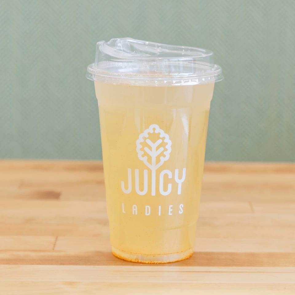 Clear plastic cup of yellow juice with a domed lid and Juicy Ladies logo on a wooden table.